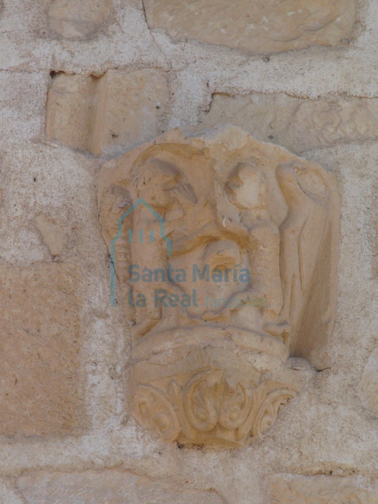 Capitel reutilizado en el muro norte sobre la portada. Un pájaro, un árbol y dos figuras humanas que parecen luchar